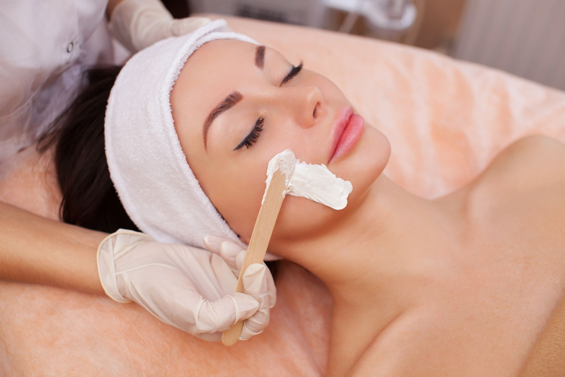 Gloved hand applies anti-aging facial treatment to woman’s face with a wooden stick.