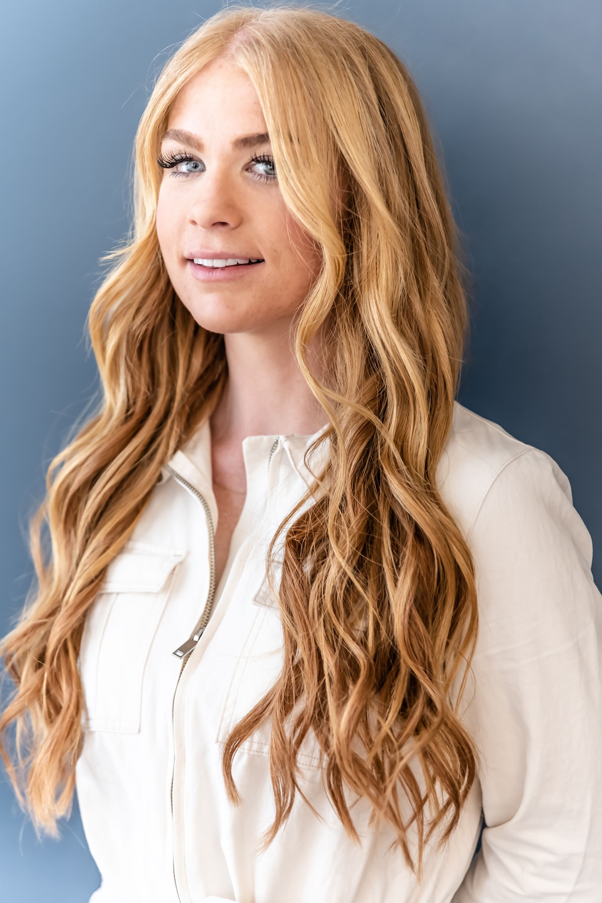 Blonde woman in a white jumpsuit smiles in front of a blue background.