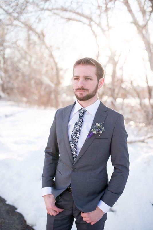 A person wearing a grey suit and a patterned tie stands outdoors in a snowy landscape with trees in the background.