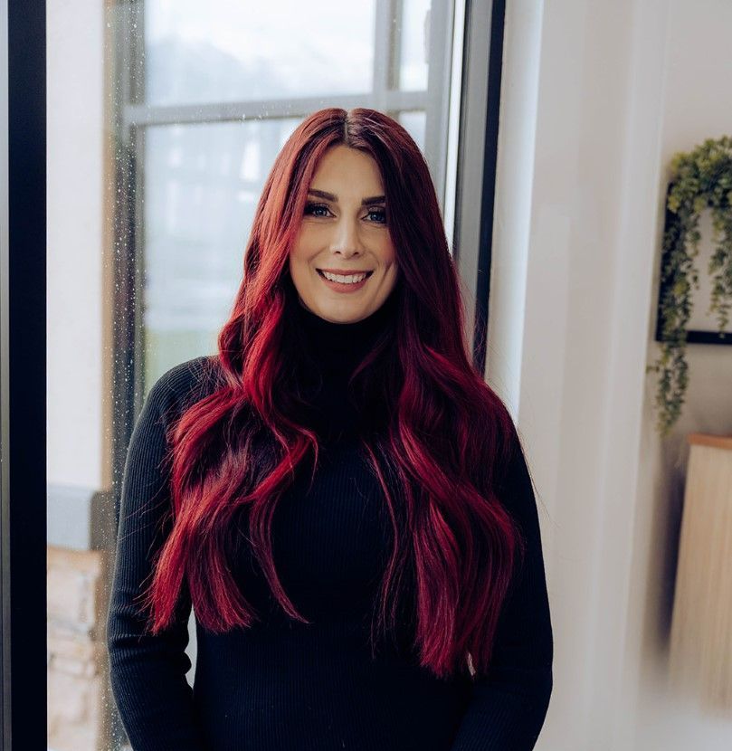 A woman with red hair is standing in front of a window and smiling.