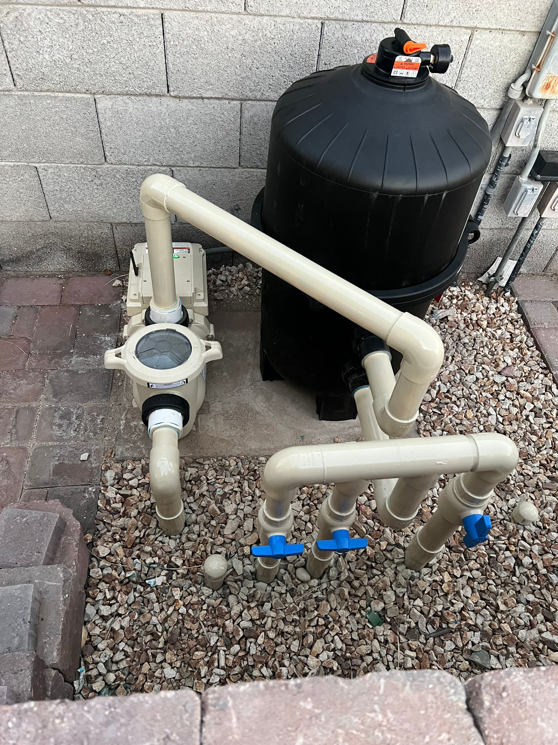 Pool filter system with beige pipes and black tank on gravel near a cinder block wall.