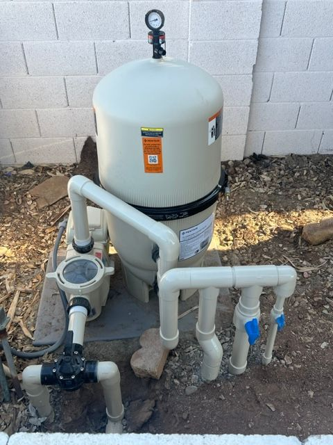 Tan pool filter system with pipes, gauge, and pump, against a white block wall.