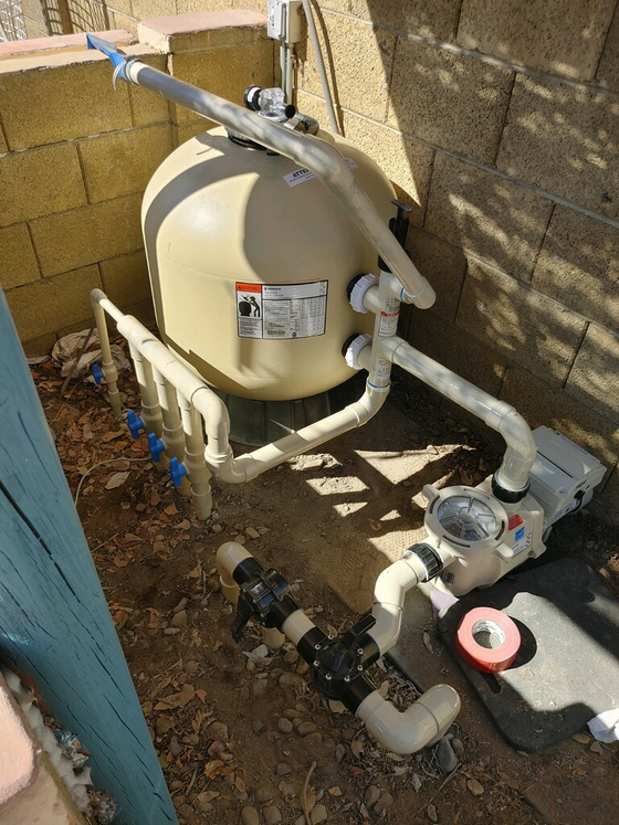Pool filter system, beige tank, connected pipes, valves, and pump, outdoor setting, partial sunlight.