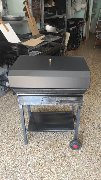 Griglia a carbone nera su supporto metallico con ripiano inferiore e una ruota a vista. All'interno, sfondo disordinato.