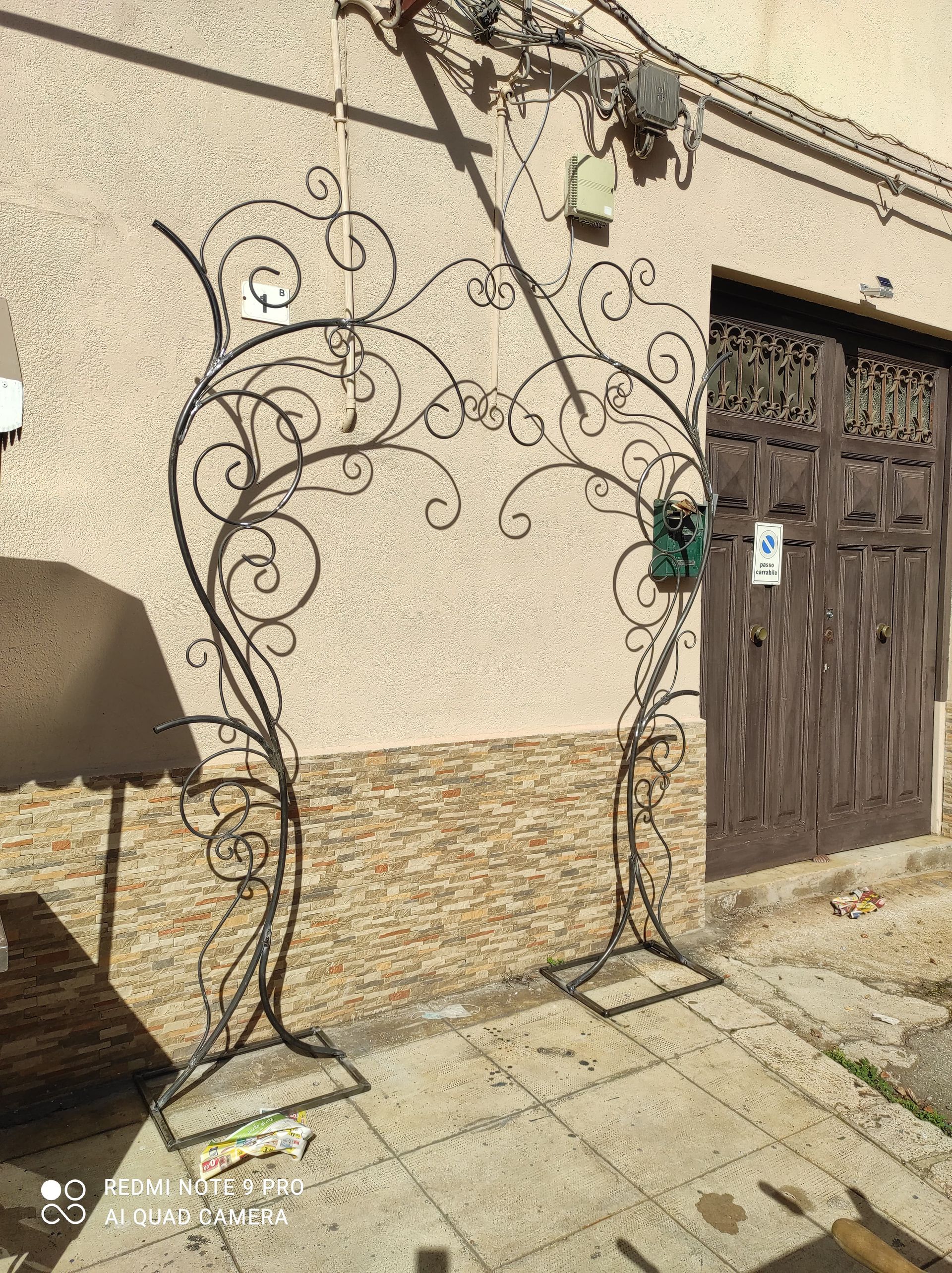 Un arco decorato in metallo nero su un marciapiede, con un muro beige e una porta marrone sullo sfondo.