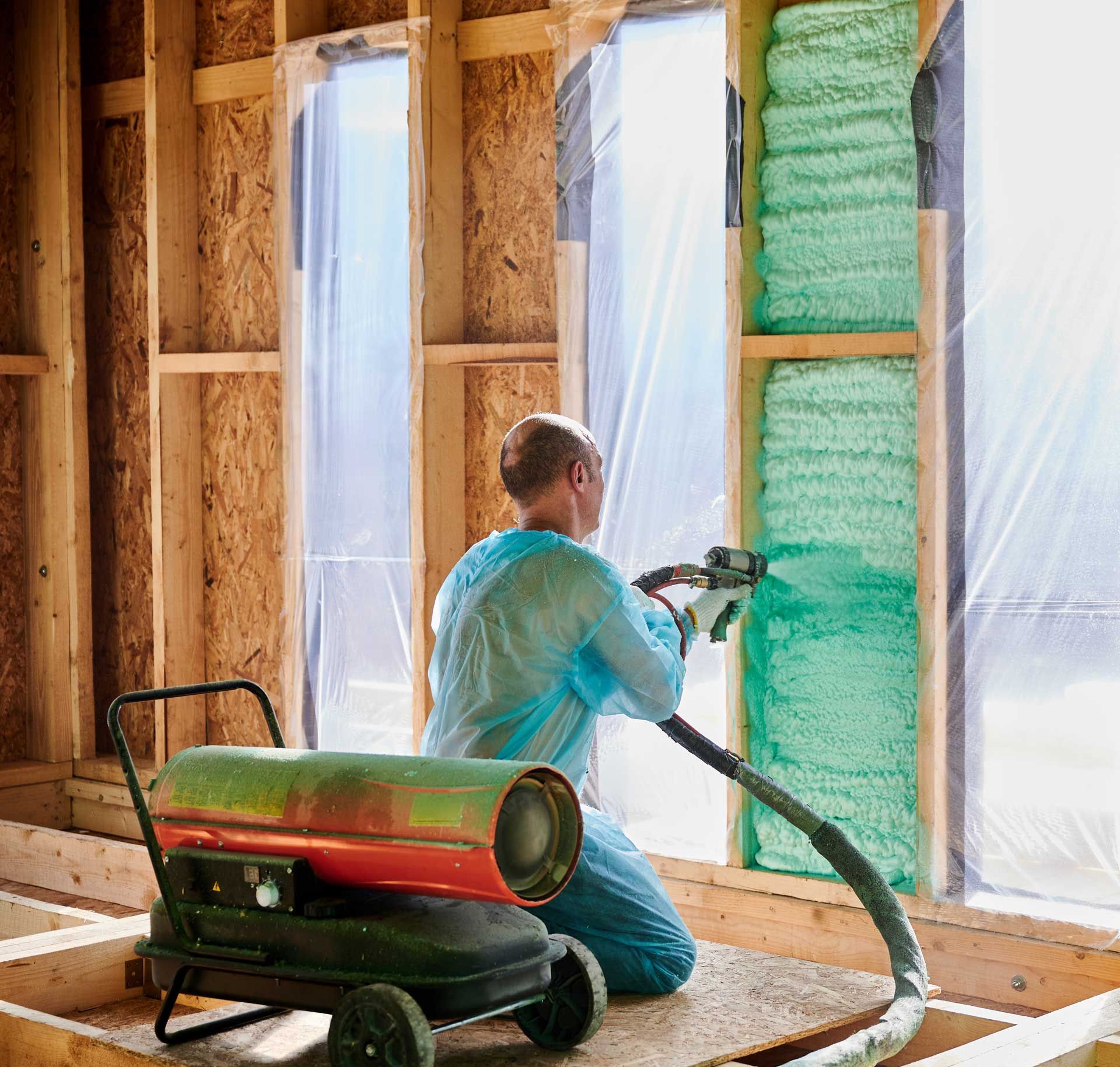 Man spraying insulation into a wall cavity with a green foam; equipment in foreground.