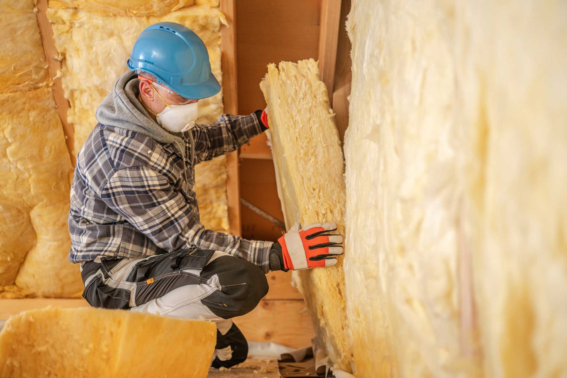 Man installing insulation in a wall, wearing a hard hat, respirator, and gloves.