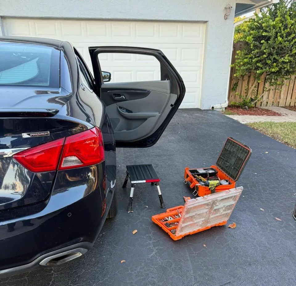 Black car in driveway with open rear door beside a toolbox and orange tool chest near a garage