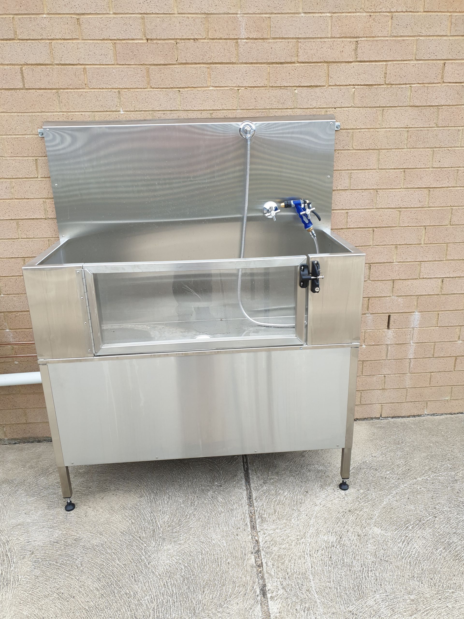 A stainless steel dog bath tub is sitting on the sidewalk in front of a brick wall.