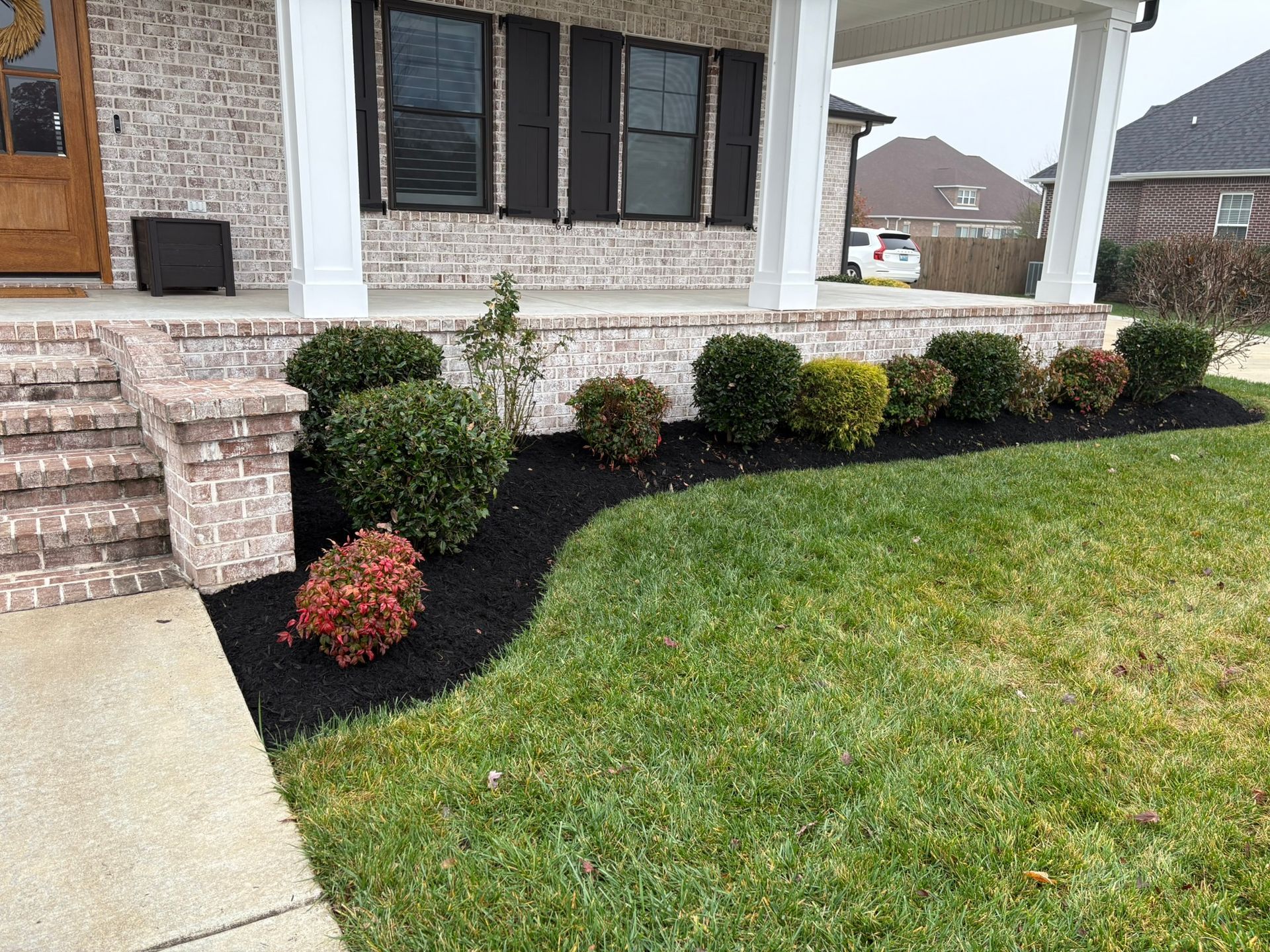 Front porch with trimmed shrubs, black mulch bed, and green lawn beside a stone-and-siding house.