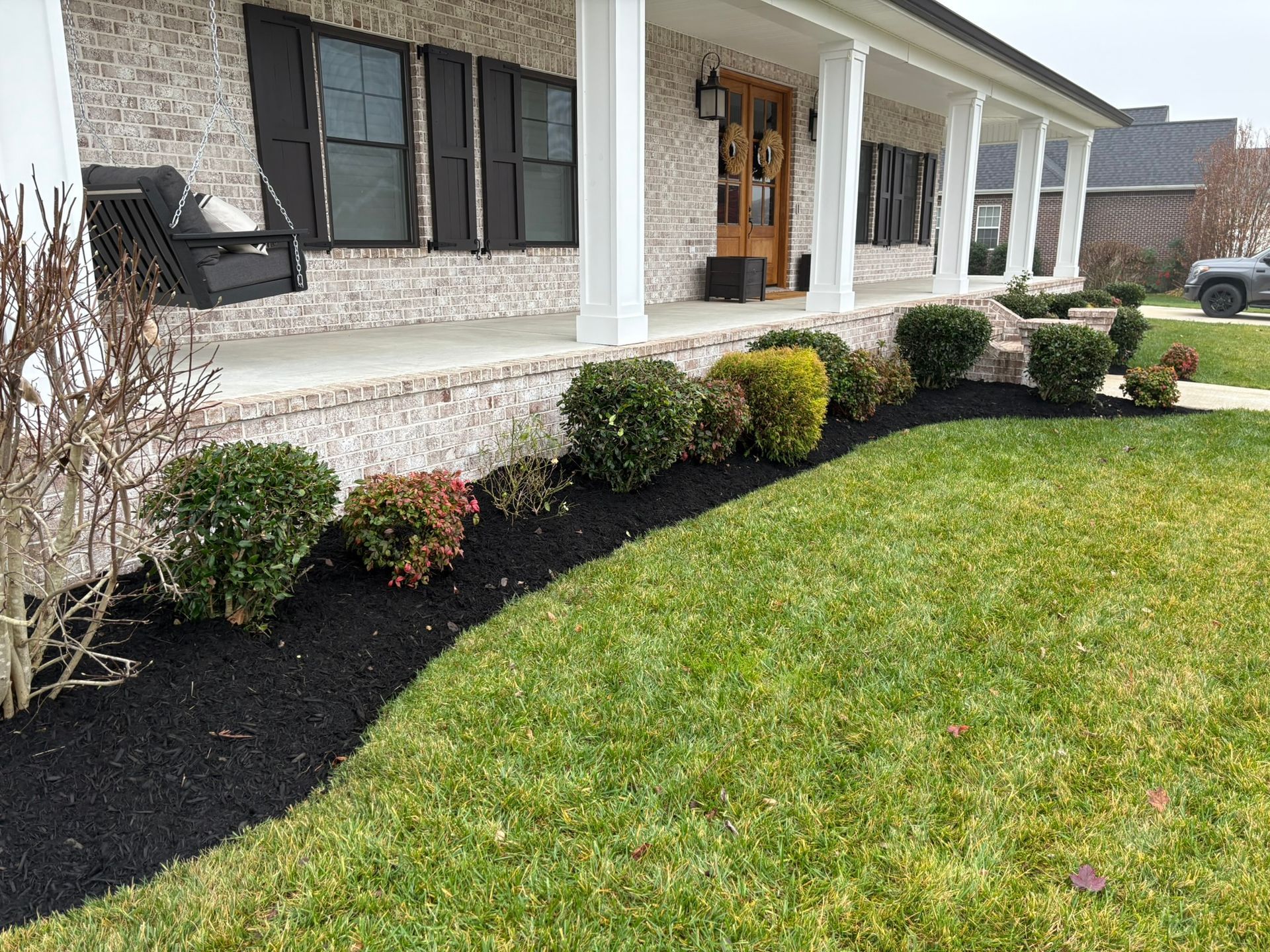 Front yard with manicured shrubs, black mulch, green lawn, and a light-colored house with a porch.