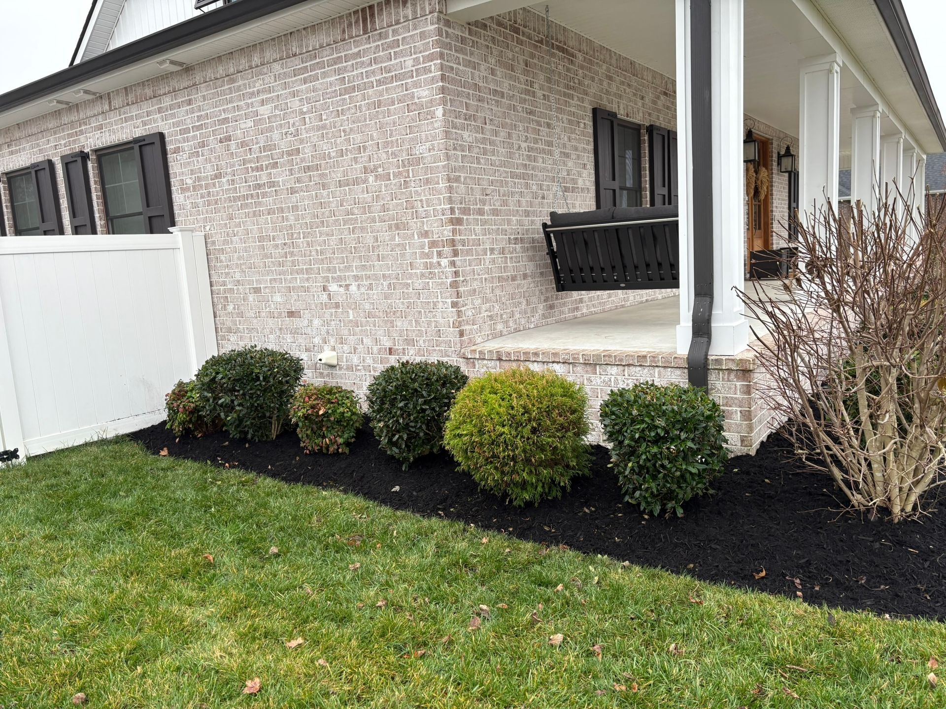 Suburban house exterior with brick walls, white porch columns, and trimmed shrubs along a black mulch bed