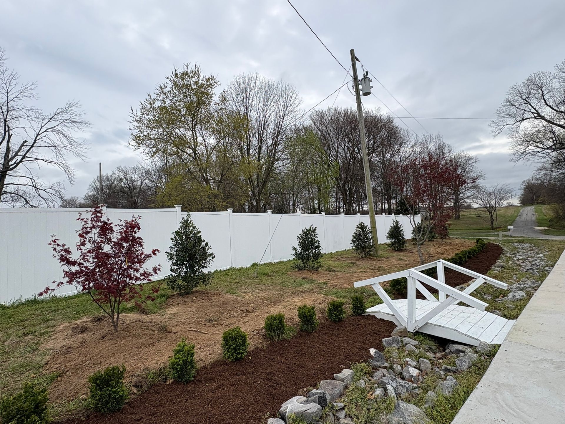 Landscaped yard with white fence, young shrubs, and a white wooden bridge over a stone-lined path