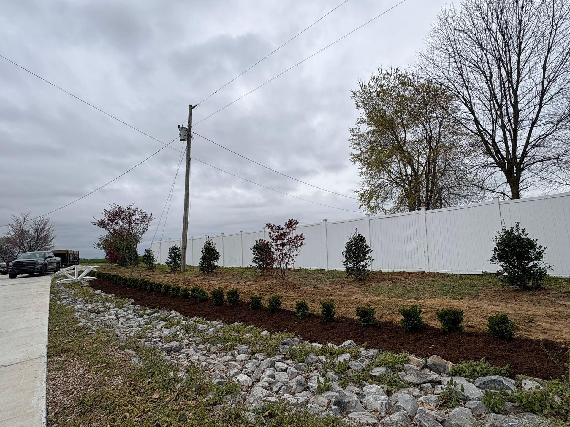 Landscaped roadside with young shrubs, stone retaining wall, white fence, and cloudy sky.