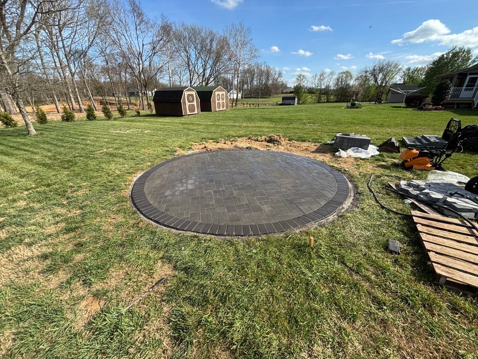 Round black trampoline or platform in a grassy yard with scattered materials and a small shed in the background
