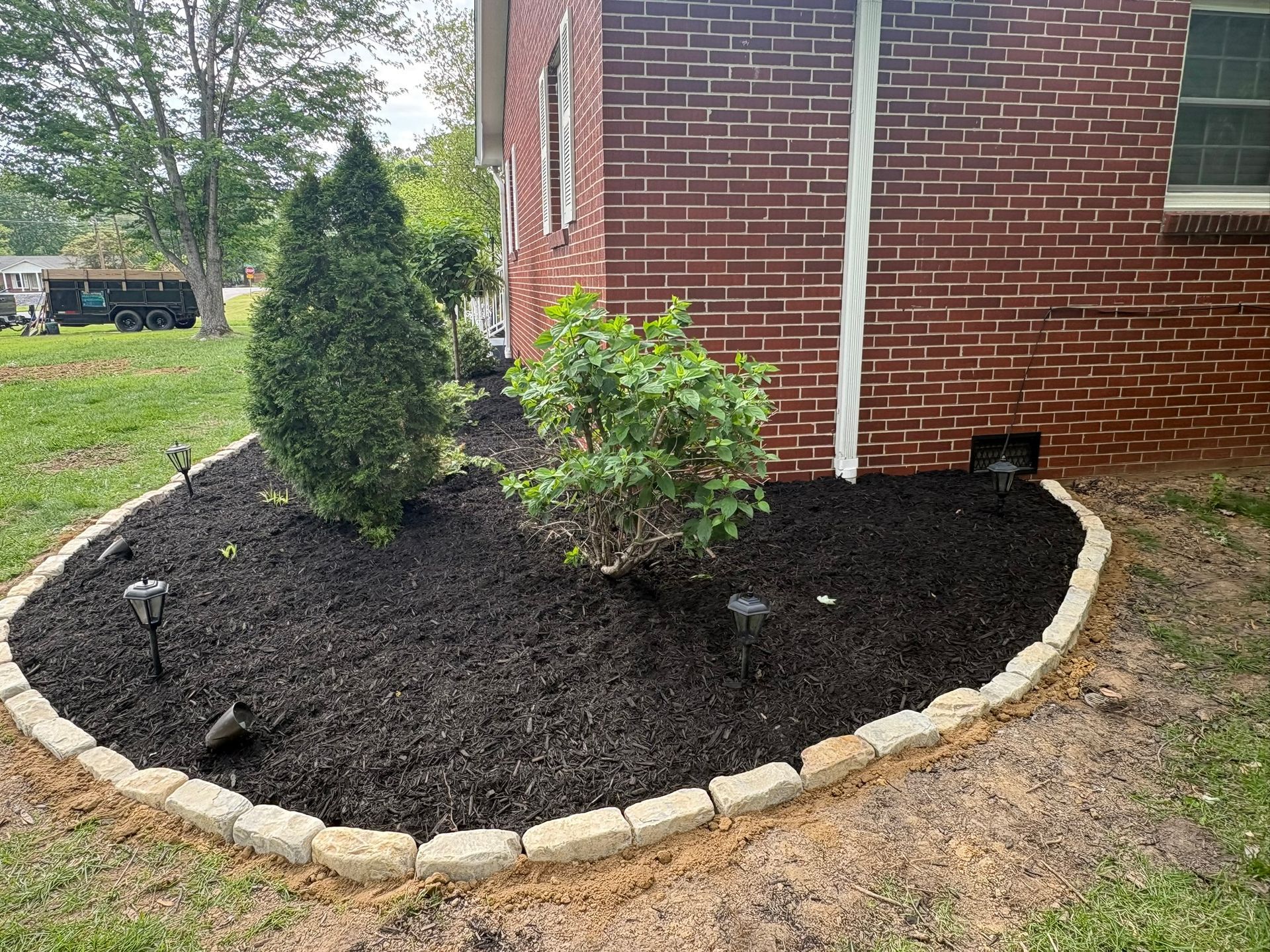 Mulched flower bed with two shrubs beside a brick house, bordered by pale stone edging