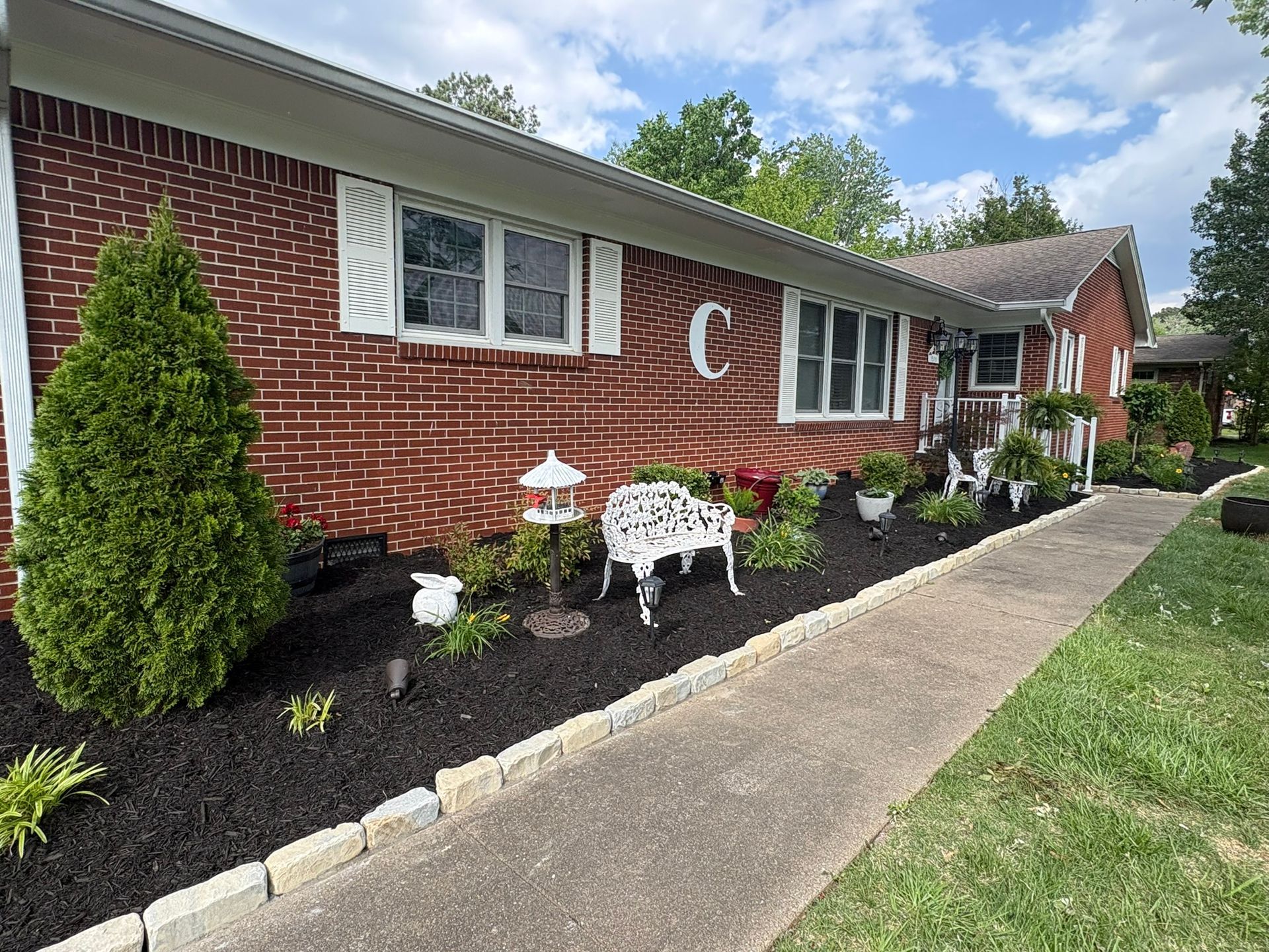 Brick house with black mulch, white garden bench, and trimmed shrubs along a sidewalk