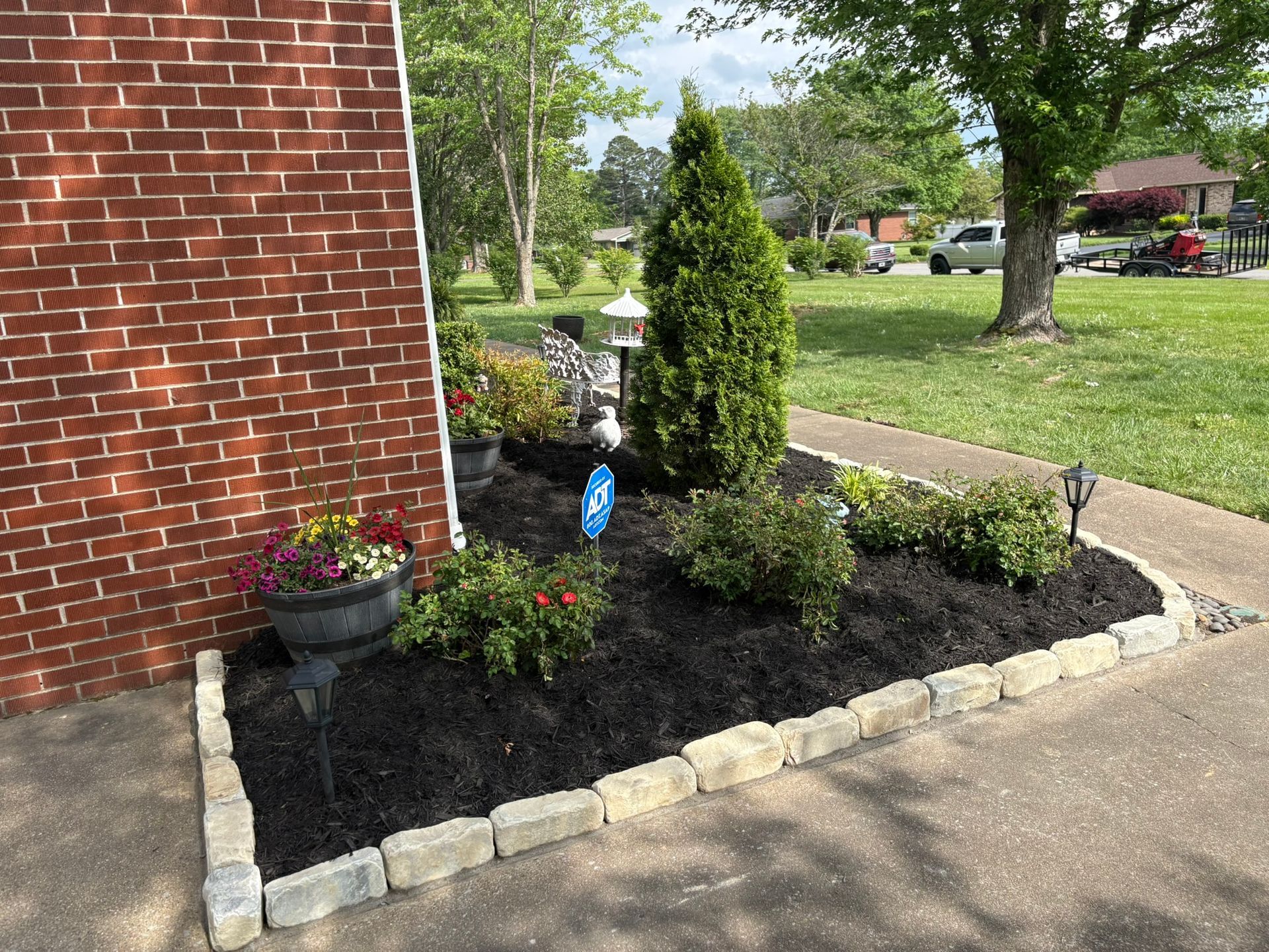 Small landscaped flower bed beside a brick wall, with dark mulch, stone border, shrubs, and flowers.