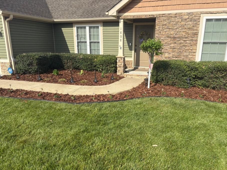 Front yard landscaping with green lawn, curved walkway, shrubs, and a stone-and-siding house entrance.