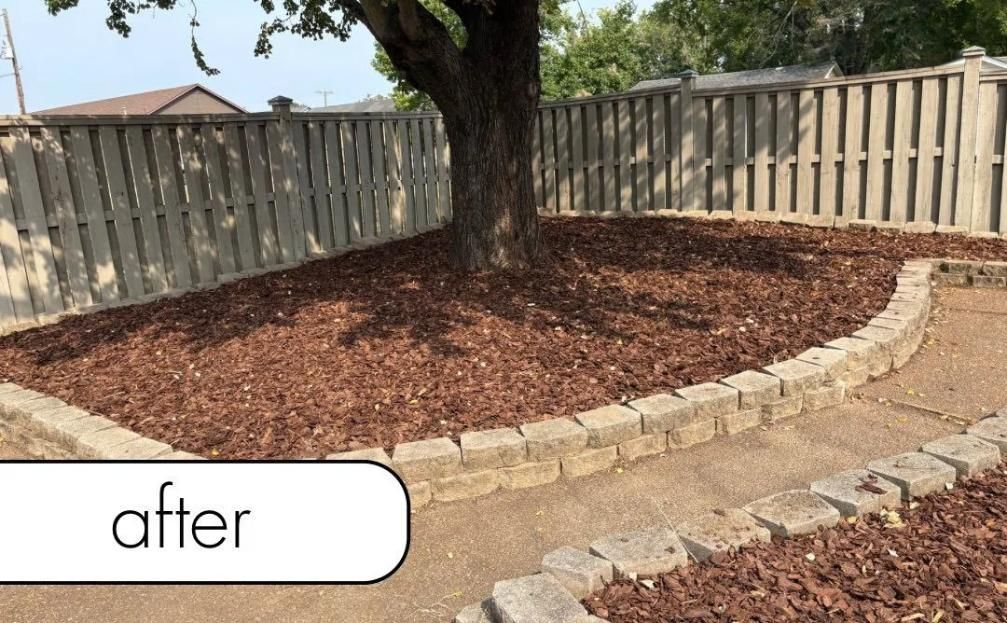 Mulched backyard garden bed around a large tree beside a wooden fence and brick edging