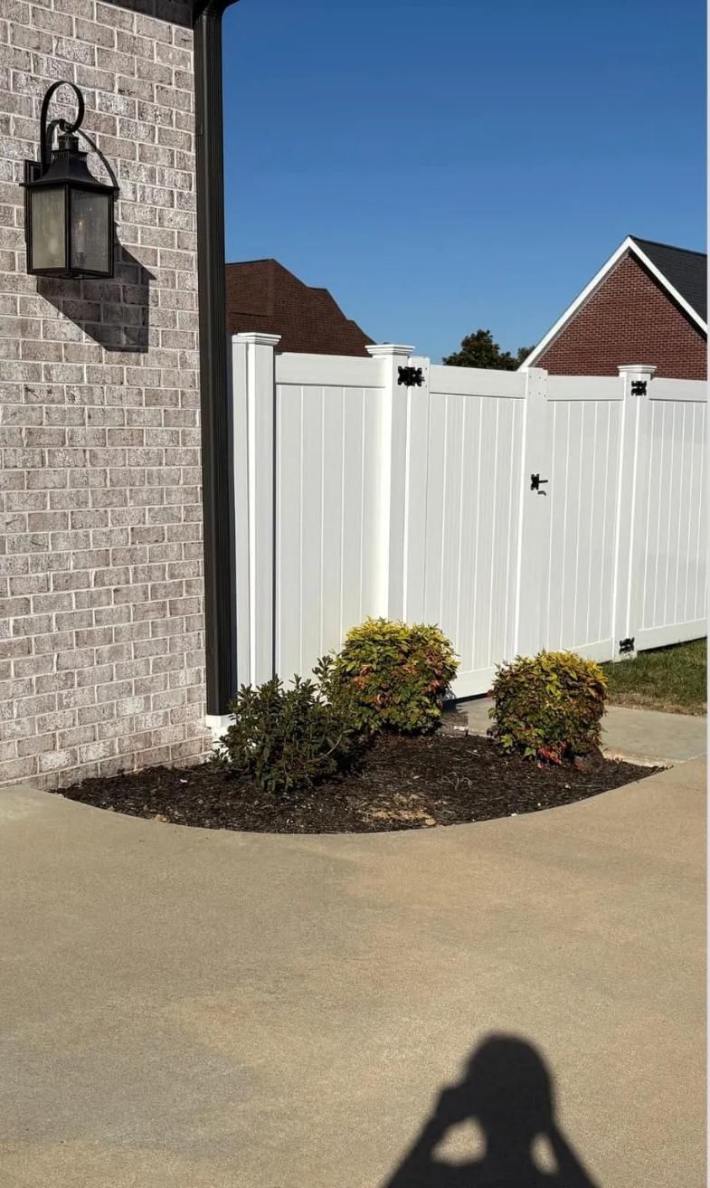 White fence beside a stone house, with a small shrub and patio under a clear blue sky.