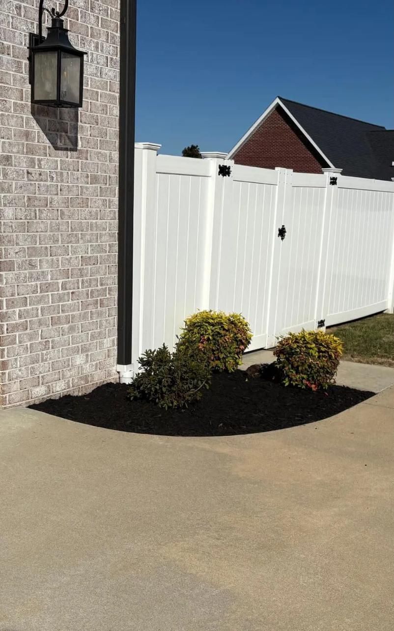 White fence beside a brick house, with trimmed shrubs and a concrete walkway in sunlight