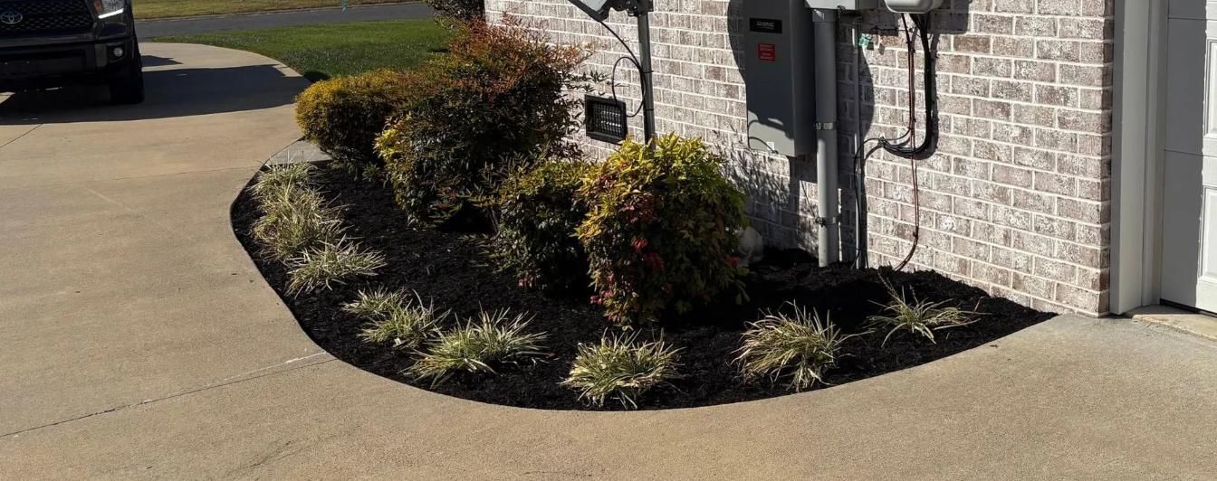 Landscaped shrub bed with mulch beside a concrete driveway and stone house wall