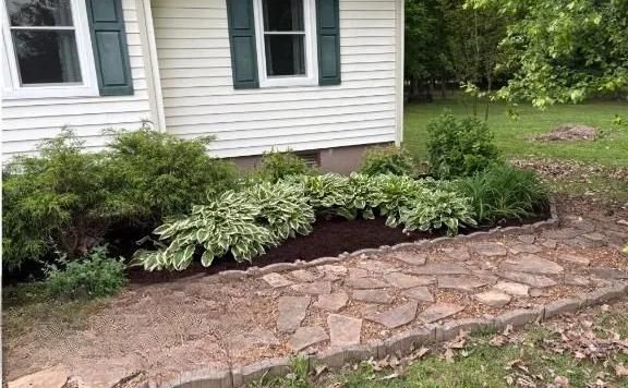 Stone walkway beside a house, bordered by green shrubs and dark mulch.