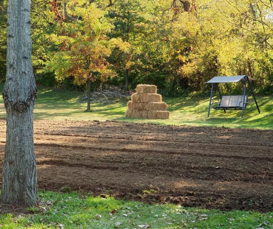 Hay bales in a freshly tilled field with autumn trees and a swing bench in the background.
