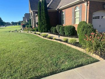Suburban brick house with a neatly trimmed lawn, shrubs, and a concrete driveway in bright sunlight