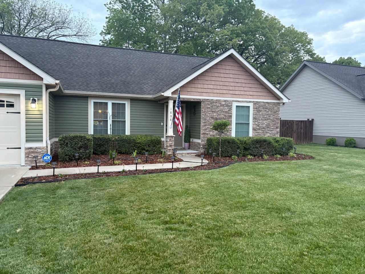 Front yard landscaping with shrubs, mulch beds, and a concrete walkway beside a house
