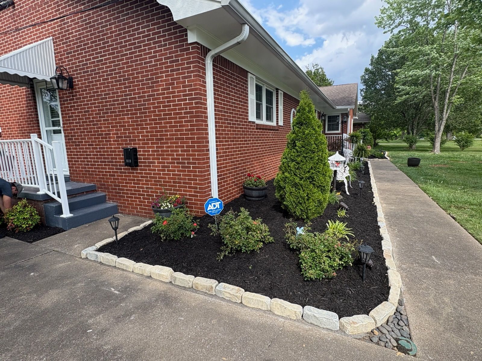 Brick house with a small landscaped bed, black mulch, shrubs, and a concrete walkway along the side.