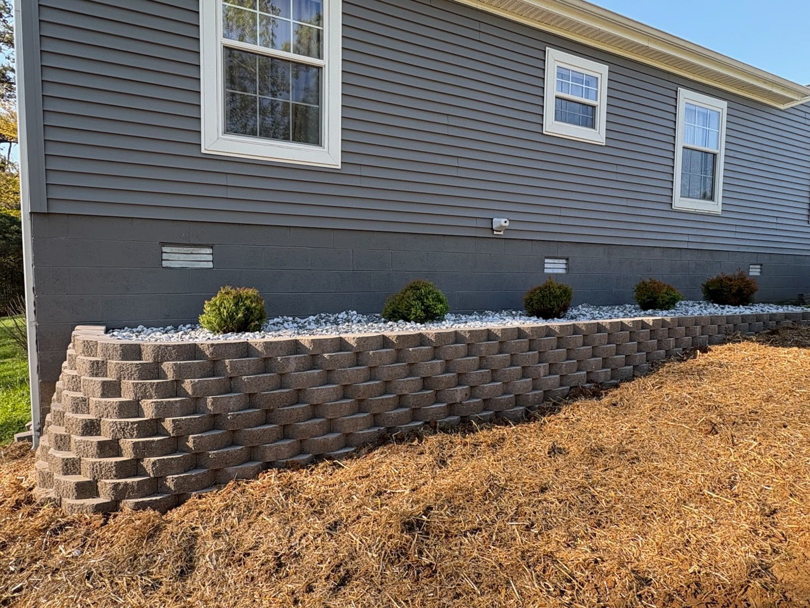 Gray house exterior with a tiered retaining wall, small shrubs, and mulch-covered landscaping