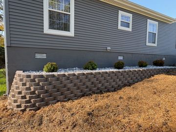 Brick retaining wall on a grassy roadside slope beside suburban houses, with a tractor and loader nearby.
