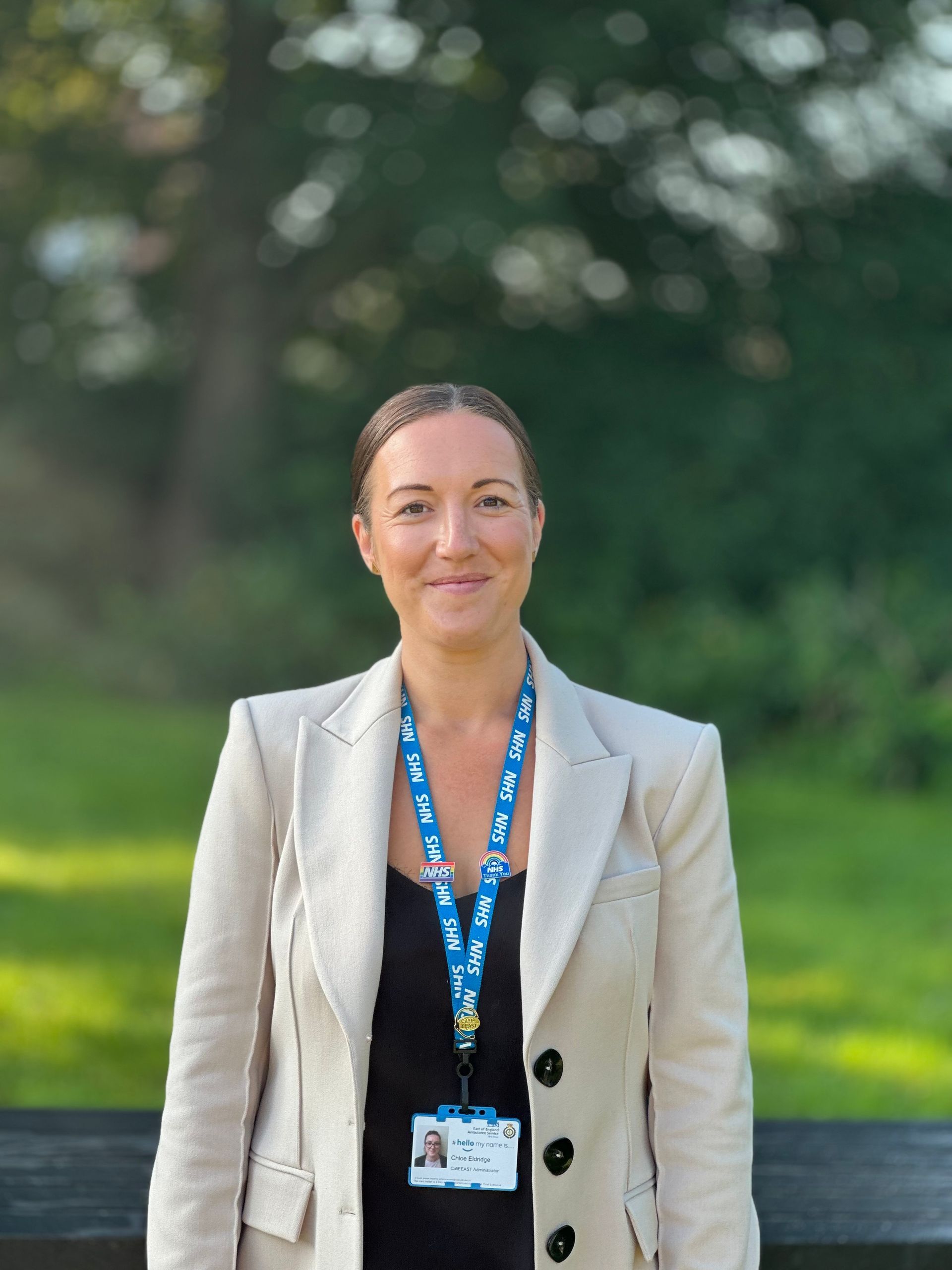 A lady with brown hair tied up. She is wearing a black top and neutral coloured blazer. She is smiling, is wearing her NHS lanyard and has a blurred background. 
