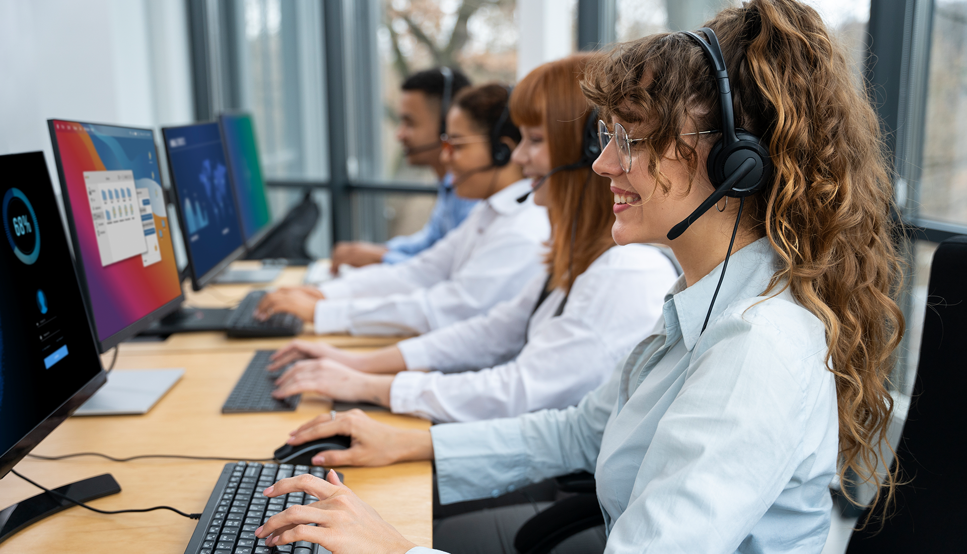 A group of people wearing headsets are sitting in front of computer monitors in a call center.