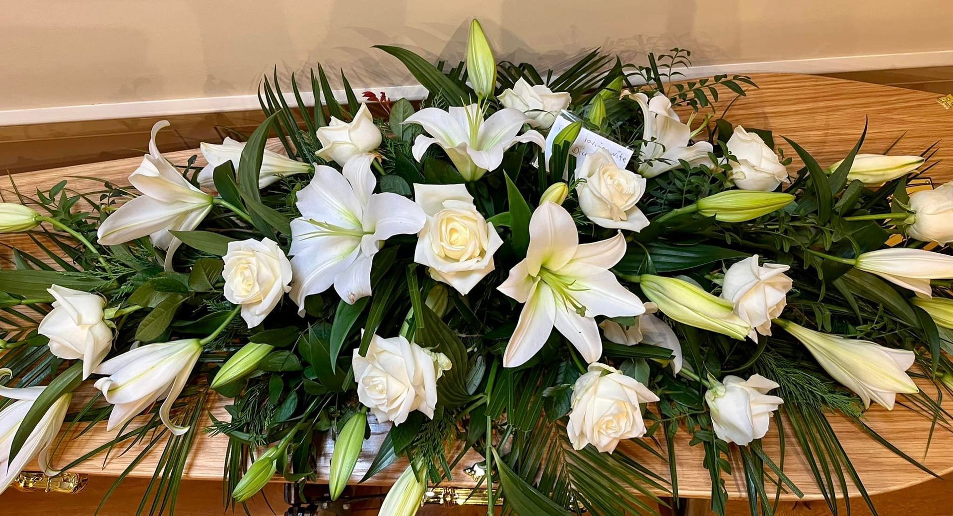 A coffin covered in white flowers and greenery is sitting on a wooden table.