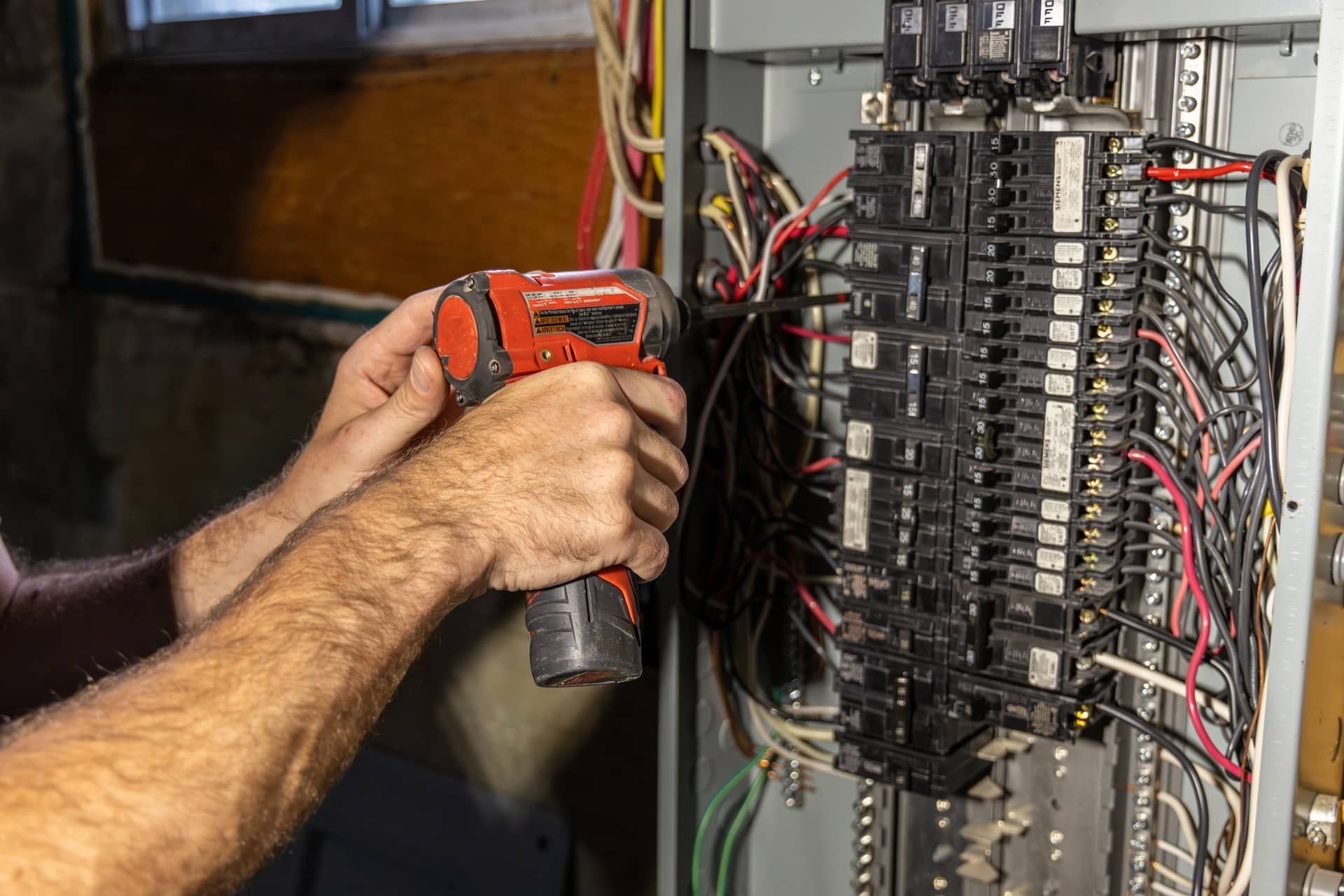 Electrician using a drill to work on an electrical control panel with wires and circuit breakers.