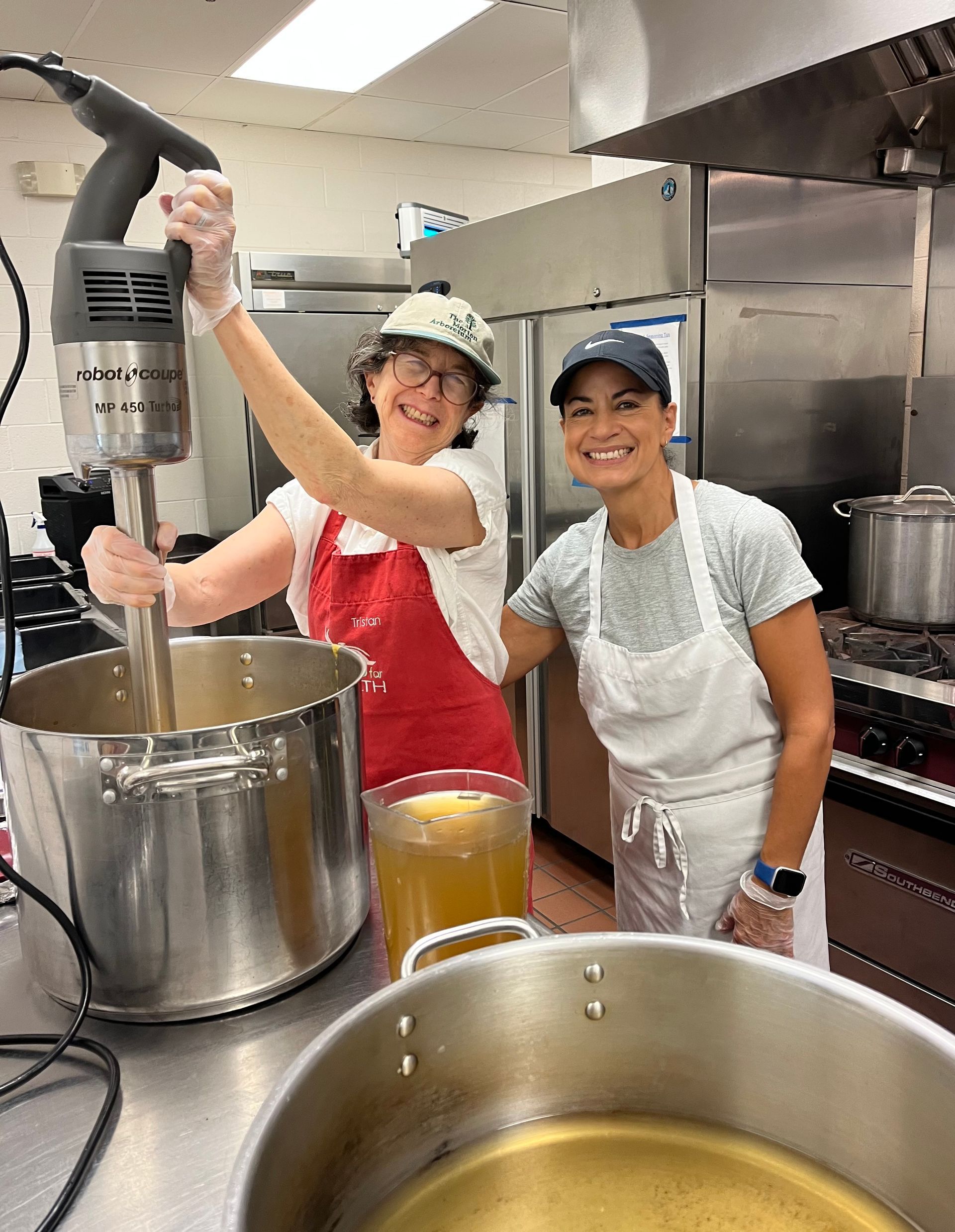 Two kitchen staff stirring large pots in a commercial kitchen, smiling at the camera.