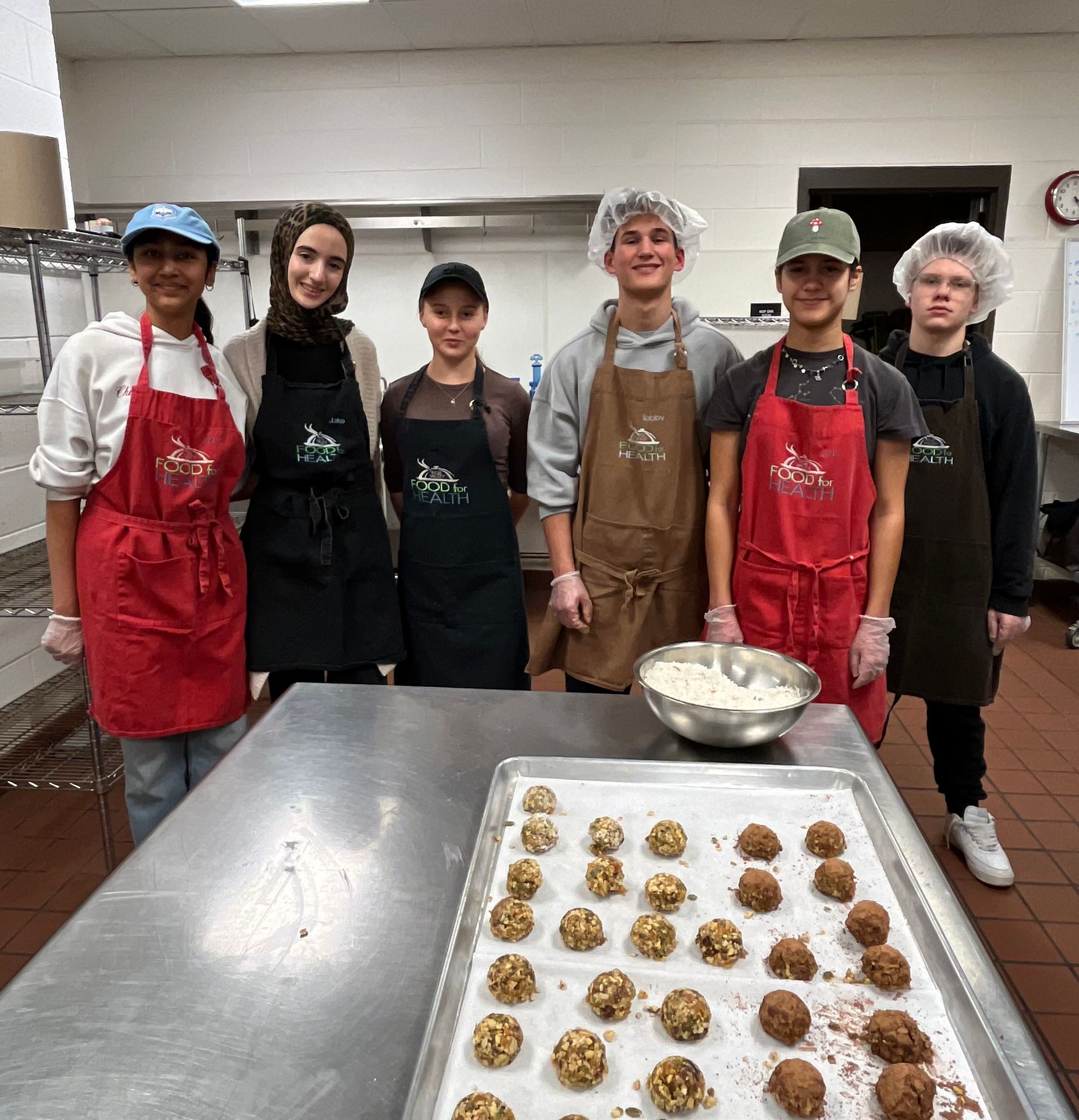 Five people in aprons stand behind trays of cookie dough in a commercial kitchen.