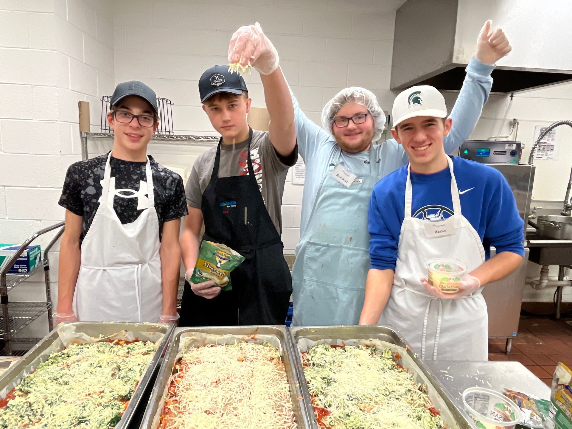Four volunteers in aprons and gloves stand behind trays of prepared food in a kitchen.