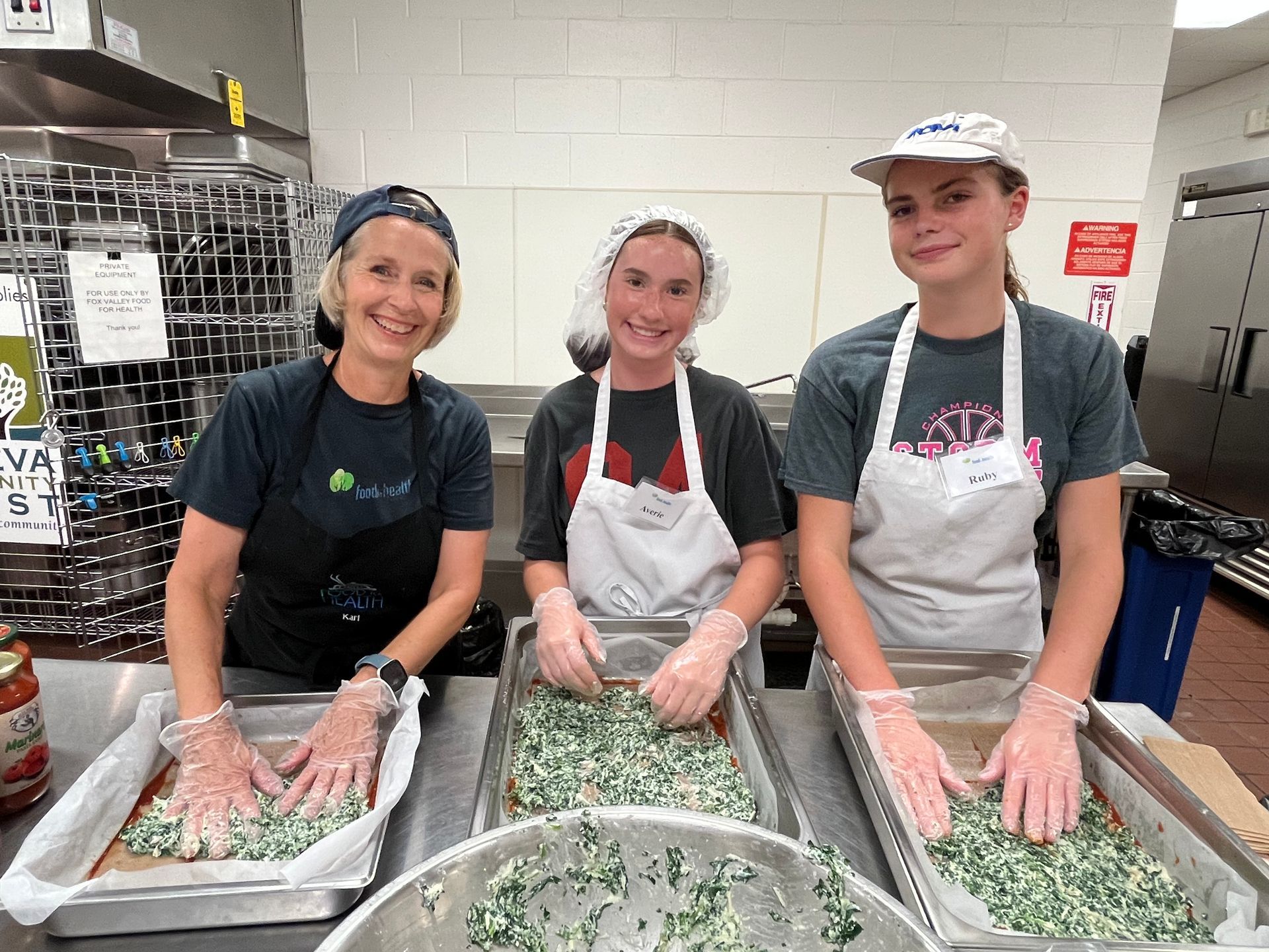 Three people in aprons mixing ingredients in large trays in a kitchen.