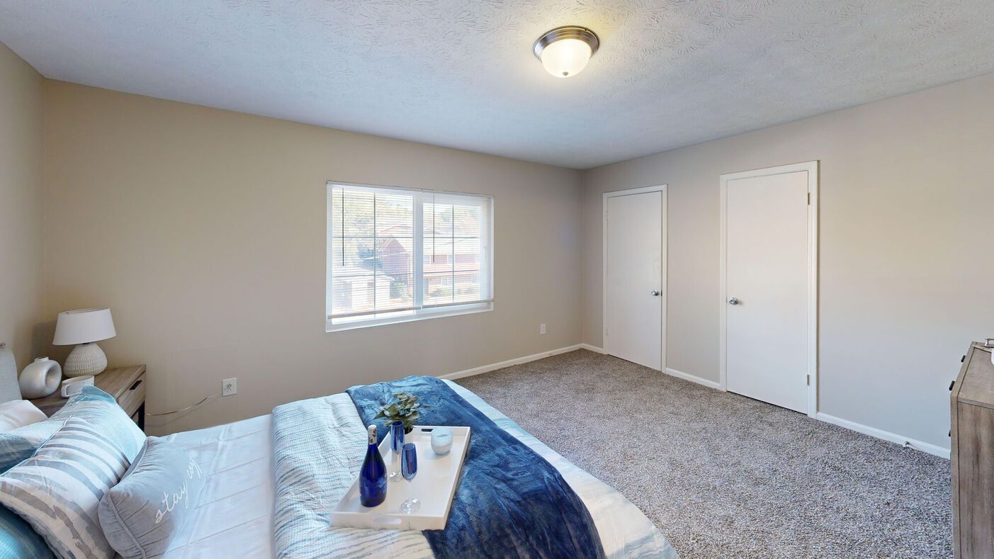 Bedroom with bed, window, two doors, nightstand, lamp, and a tray. Beige walls and carpet.