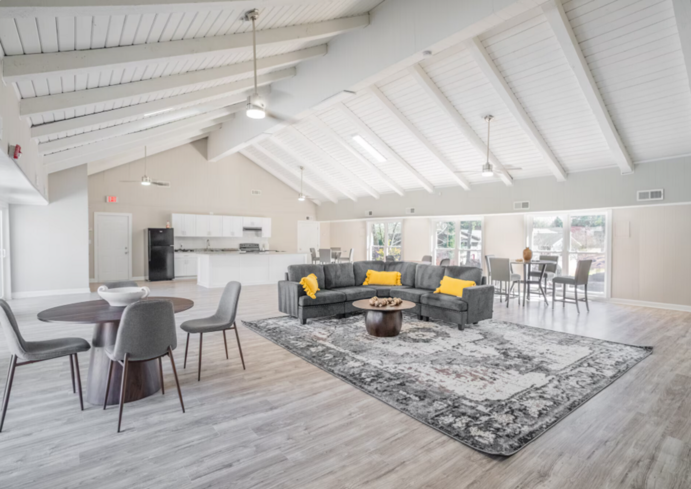 Spacious, modern interior with grey sectional, round tables, and a high, white-beamed ceiling.
