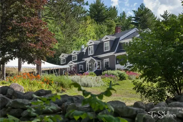 Row of dark-blue houses with white trim beside a colorful garden and trees under a partly cloudy sky