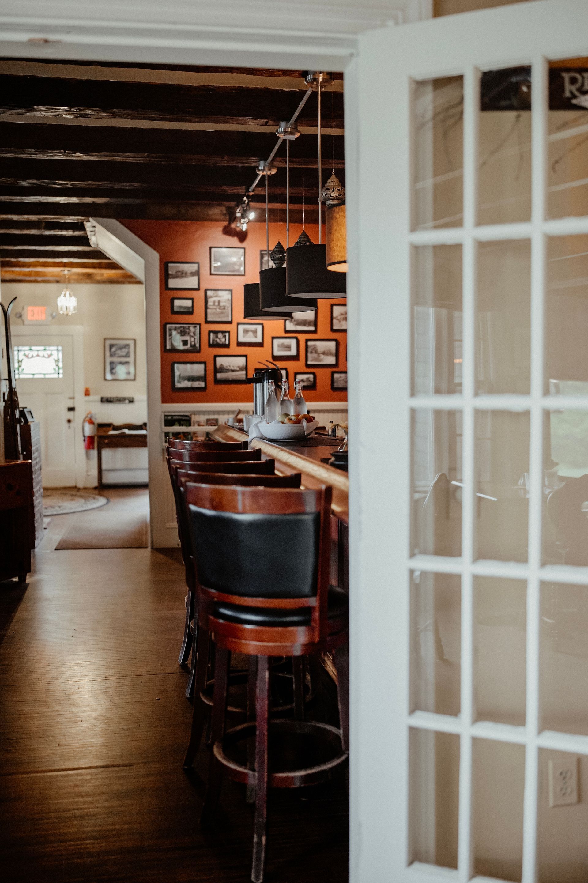 Cozy bar interior with wooden stools, framed photos on orange wall, seen through a white-paned doorway