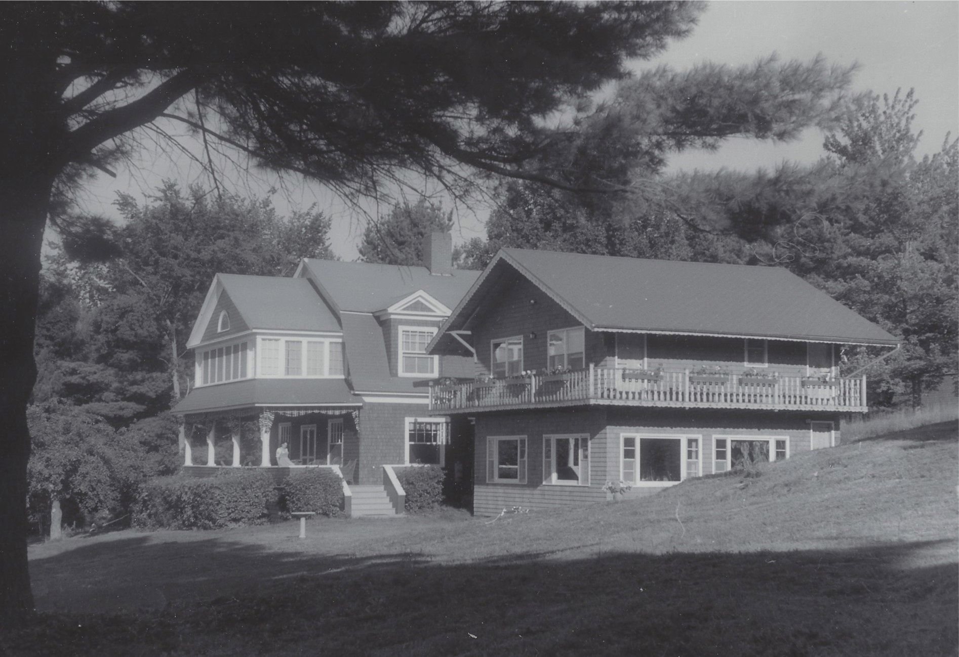 Large house with multiple gables on a grassy hillside, framed by trees in black and white.