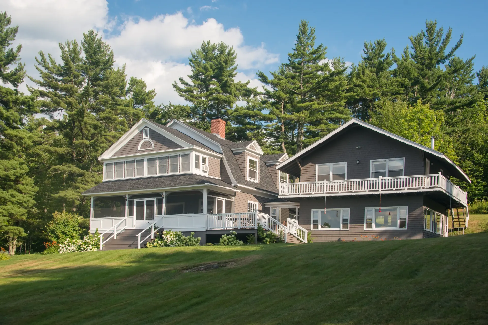 Two houses on a green lawn beside tall trees under a partly cloudy sky