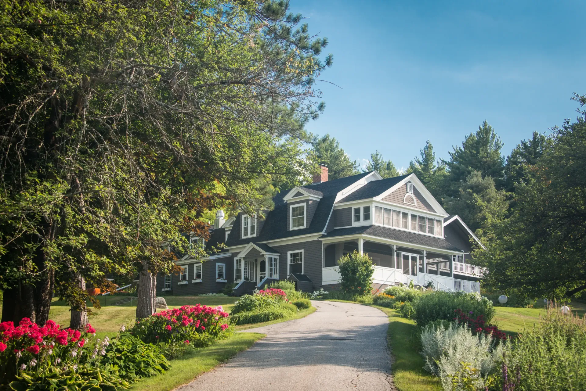 Large gray house with white trim beside a curved path, surrounded by trees, flowers, and greenery under a blue sky