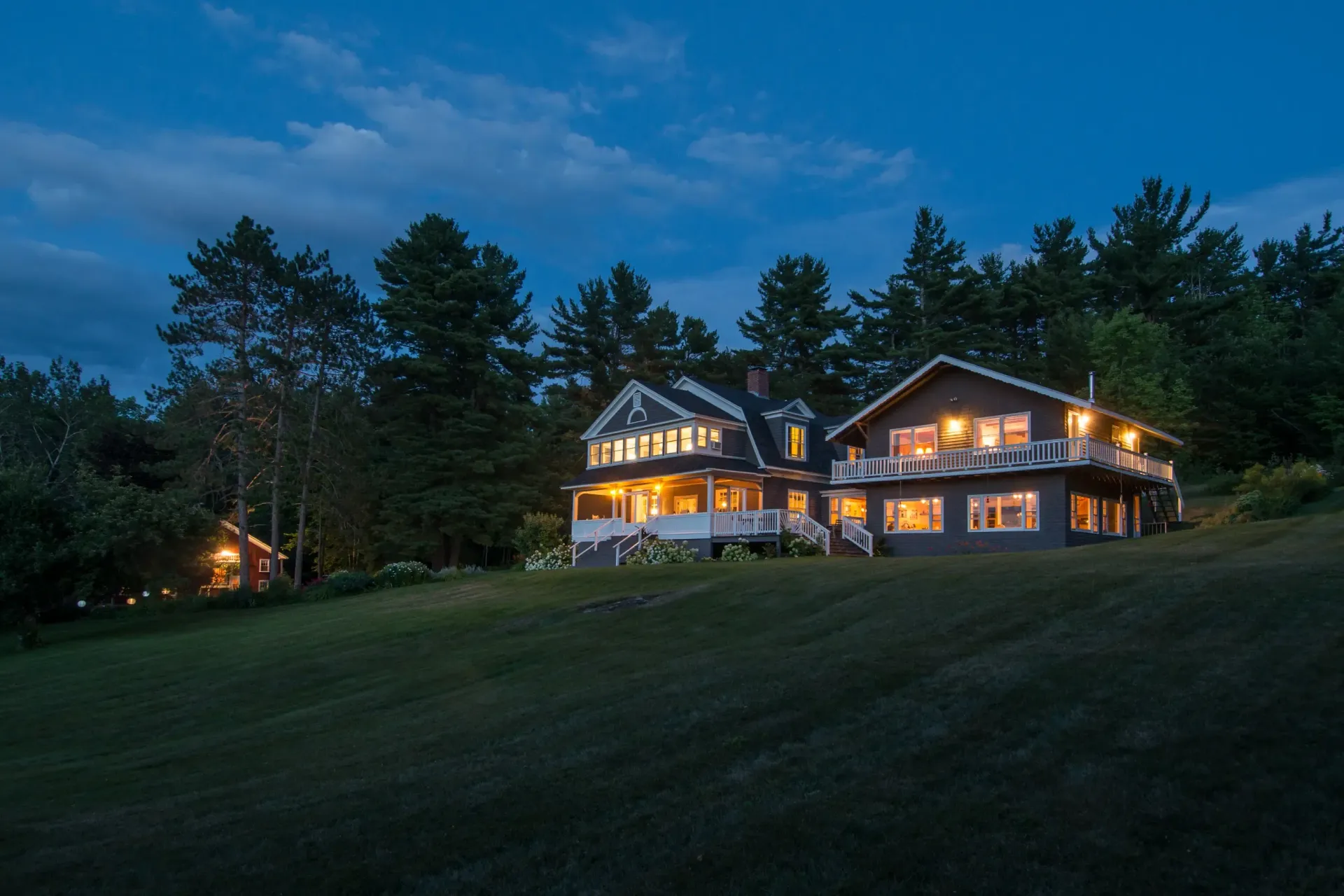 Lit lakeside lodge at dusk on a grassy hillside, with trees silhouetted against a blue evening sky